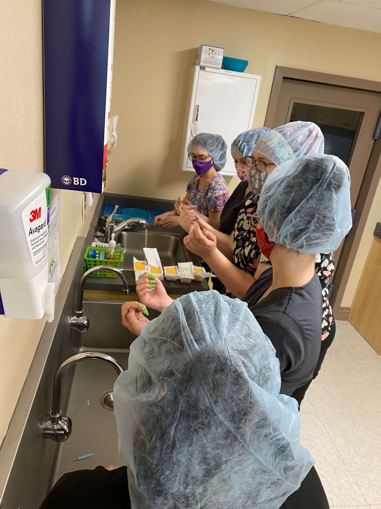 Five students wearing hair coverings, medical scrubs, and face masks, washing their hands